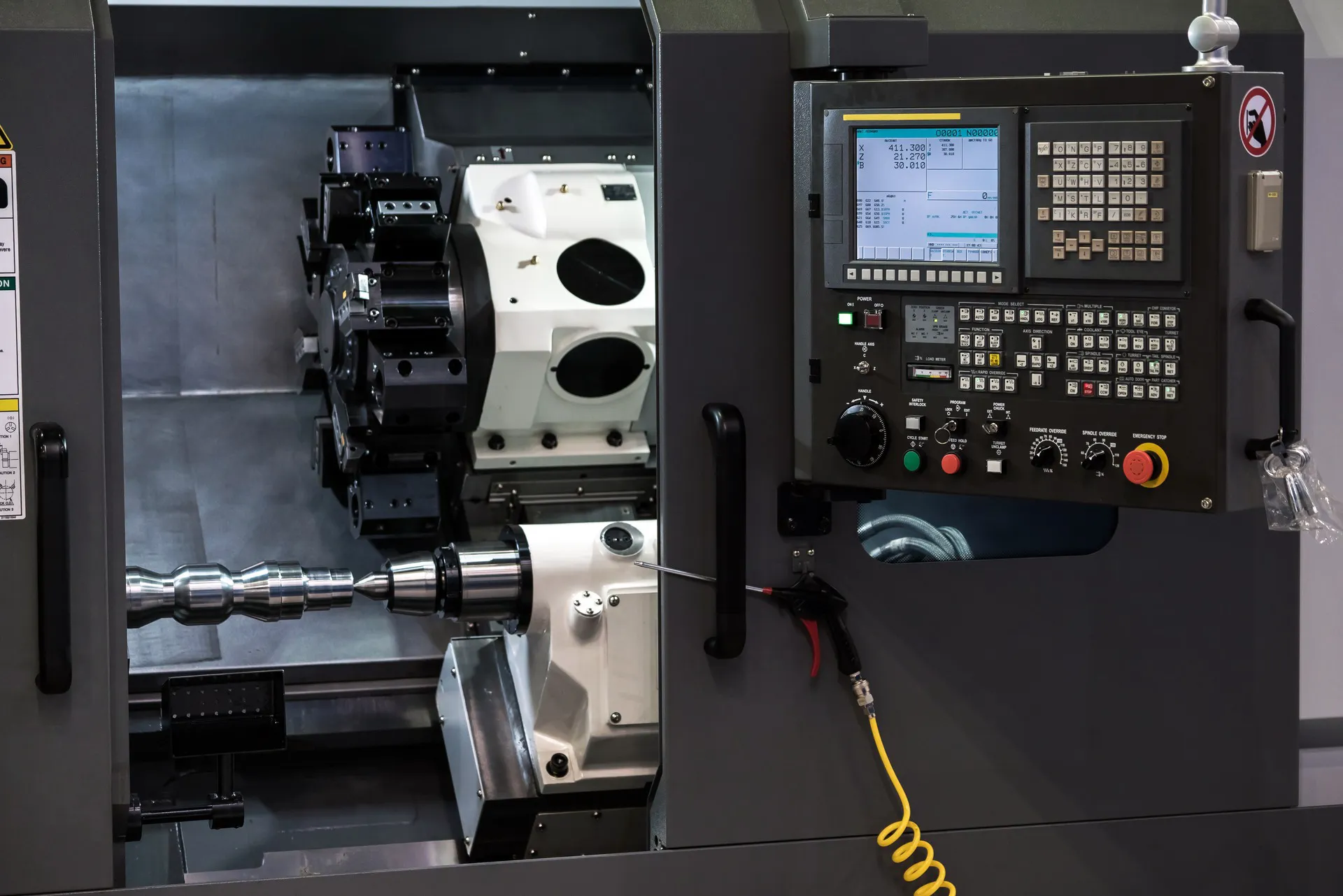A CNC gang-type turning centre — a machined metal workpiece between centres and tooling on the left, the control panel with display, keypad and emergency stop on the right